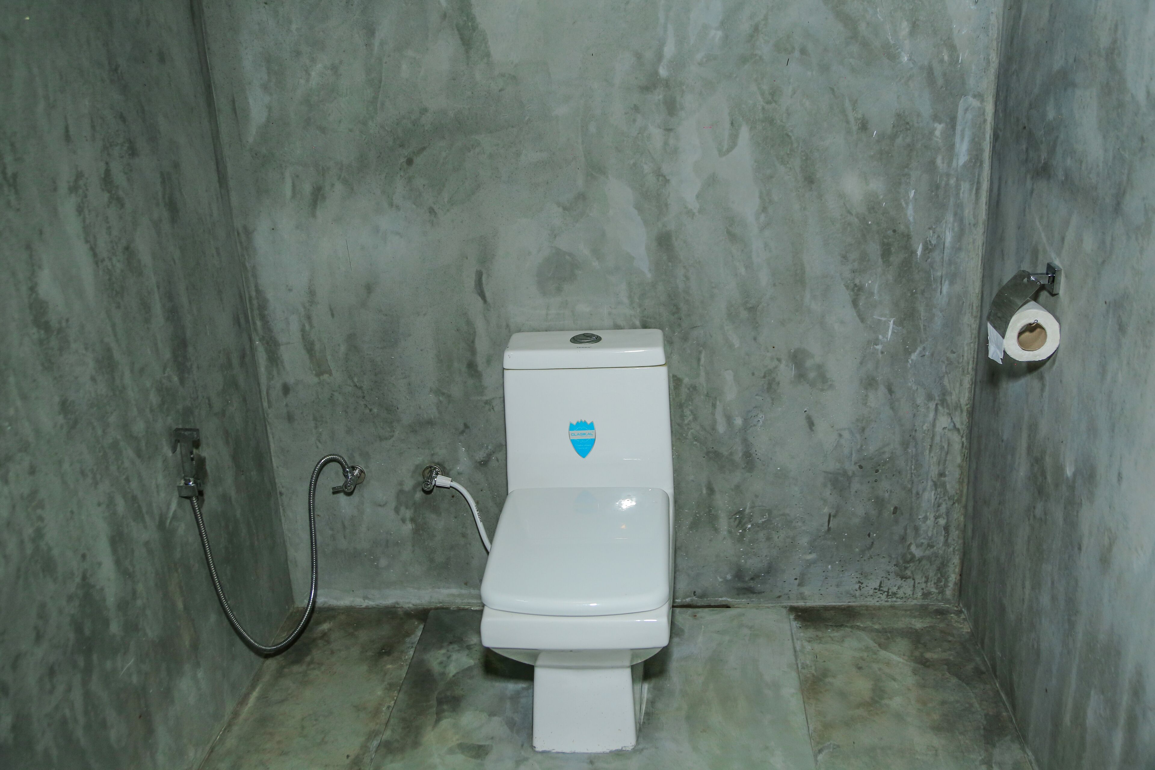 Une petite salle de bain simple, équipée d’un WC, d’une douche et d’un porte-rouleau de papier toilette fixé au mur.