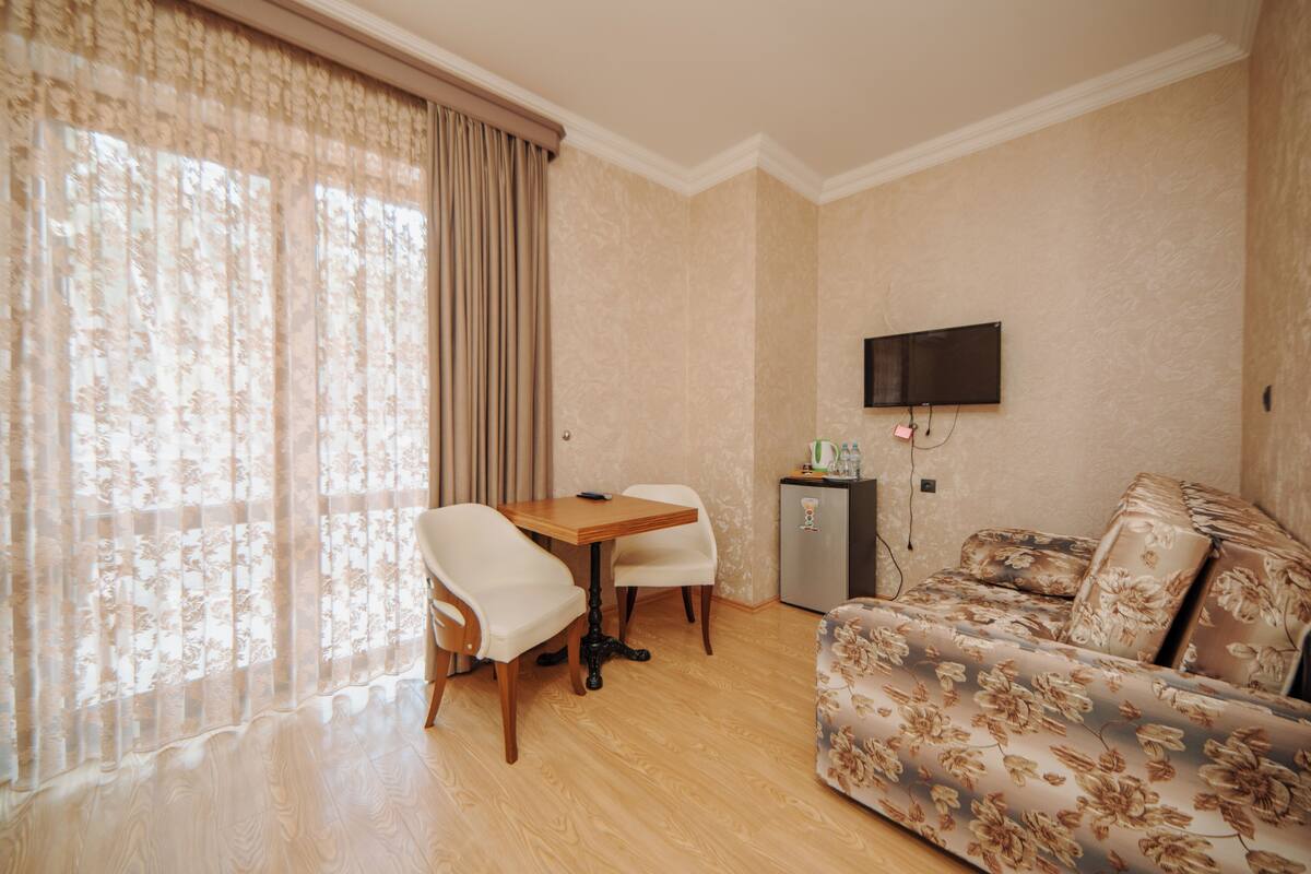 A room with a wooden floor, a table with two chairs, a sofa, a television mounted on the wall, and a small cabinet.