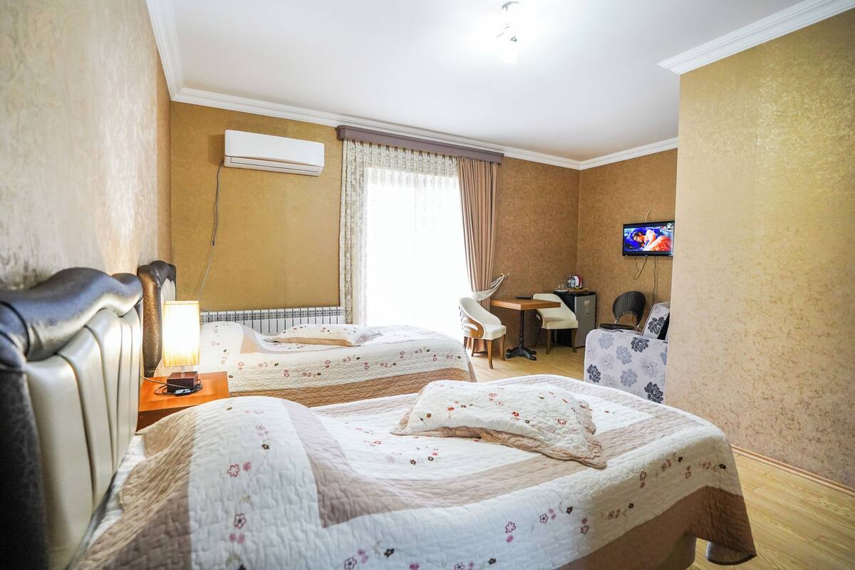 A hotel room with two beds, a desk, a TV, and an air conditioning unit.