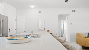 A modern kitchen with a long island, white cabinetry, and a central island with a colorful placemat. - Sunset Retreat| Pool Oasis,Walk to Beach,Billiards (Redington Beach)