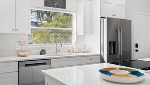 A modern kitchen with white cabinetry, a stainless steel refrigerator, and a window with blinds. - Sunset Retreat| Pool Oasis,Walk to Beach,Billiards (Redington Beach)
