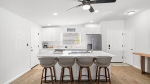 A modern kitchen with white cabinets, a central island with bar stools, a ceiling fan, and a refrigerator. - Sunset Retreat| Pool Oasis,Walk to Beach,Billiards (Redington Beach)