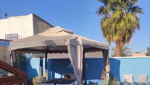 A covered outdoor seating area with a table and chairs, a large palm tree, and a blue wall in the background. - Riad L'ayinn in Marrakesh (Marrakech)