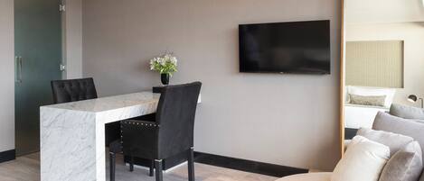 A modern hotel room with a marble desk, black upholstered chairs, a flat-screen TV, a large mirror, and a sofa with multiple cushions.