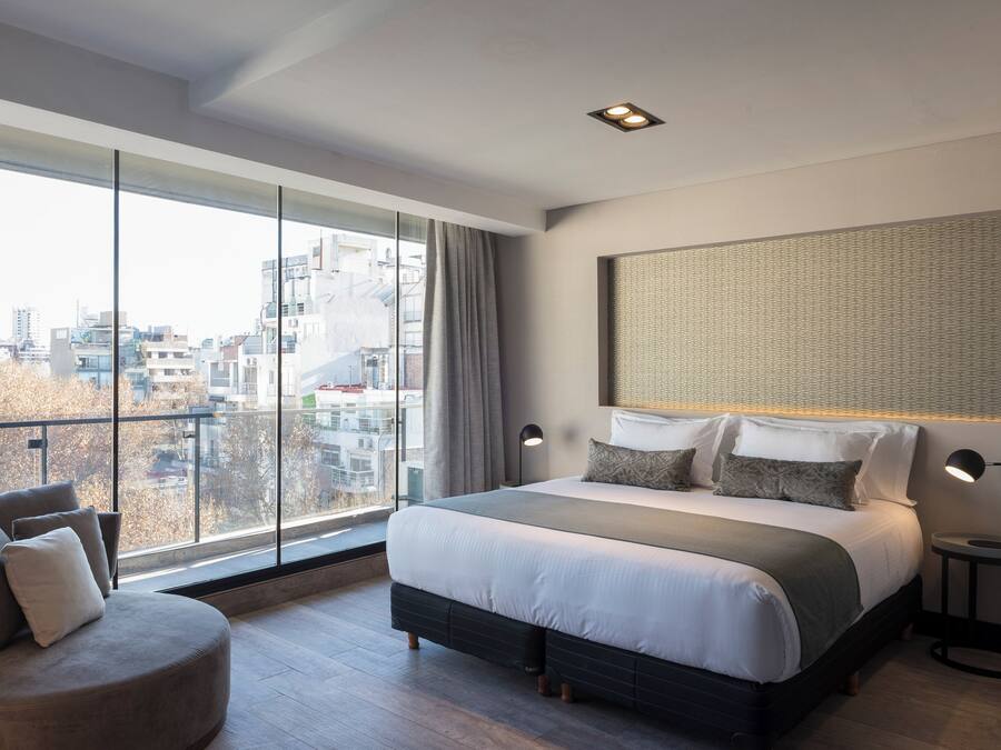 A modern hotel room with a large bed, a view of the city through floor-to-ceiling windows, a cityscape view, and a round table with a lamp.