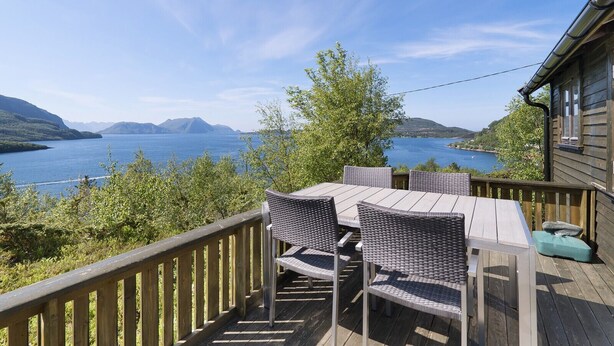 Una terraza de madera con mesa y sillas que da a un lago con montañas al fondo.