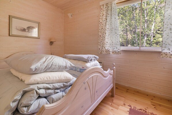 A wooden room with a bed, a framed picture on the wall, and a window with floral curtains. - Cabin - Sleeps 6 - Pets Ok - Pato - Parking (Meloy)