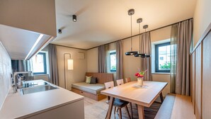 Une chambre d’hôtel moderne dotée d’une kitchenette, d’un coin repas avec une table et des chaises en bois, d’un canapé et d’une grande fenêtre avec des rideaux.