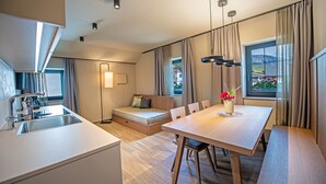 Une cuisine moderne dotée d’un évier encastré, un coin repas avec une table et des chaises en bois, un canapé avec un matelas, et une grande fenêtre agrémentée de rideaux.