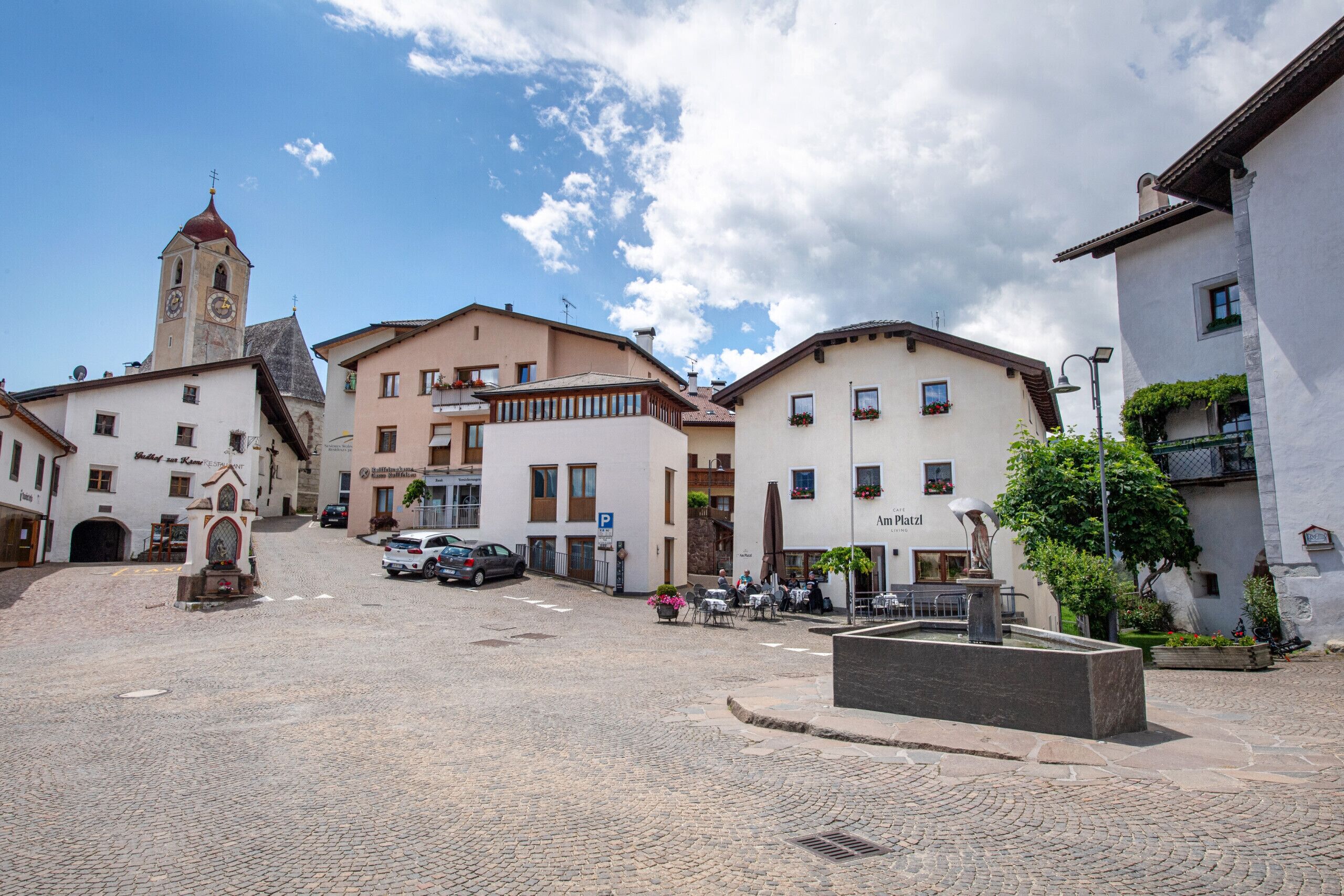 A cobblestone square with historic buildings, a clock tower, and outdoor seating areas.