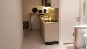 A narrow, white-walled room with a kitchenette on one side, a bed with a white headboard on the other, and a door leading to another room.