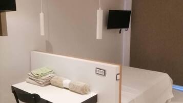 A modern bedroom with a bed, a desk, and a chair. There is a TV mounted on the wall and a power outlet on the wall near the bed.