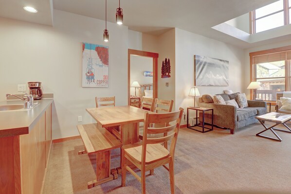 A modern living room with a wooden dining table and chairs, a sofa, and a coffee table. There is a kitchen area with a sink and a coffee maker. The room has a large window and a painting on the wall.