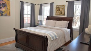 A bedroom with a wooden bed, a nightstand, a lamp, and a window with curtains. - The 87th House (Portland)