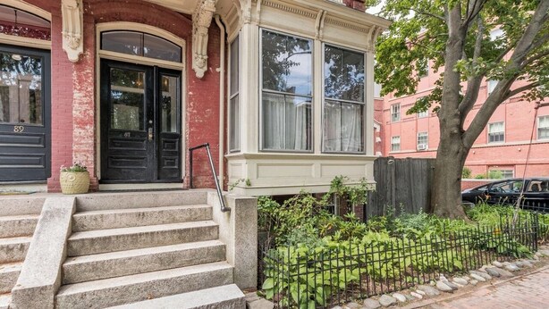 A two-story building with a black front door, a bay window, and a small garden area with a fence. - The 87th House (Portland)