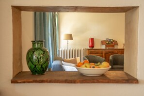 A room with a wooden shelf holding a green vase and a bowl of fruit, a radiator, a lamp, and a red vase.