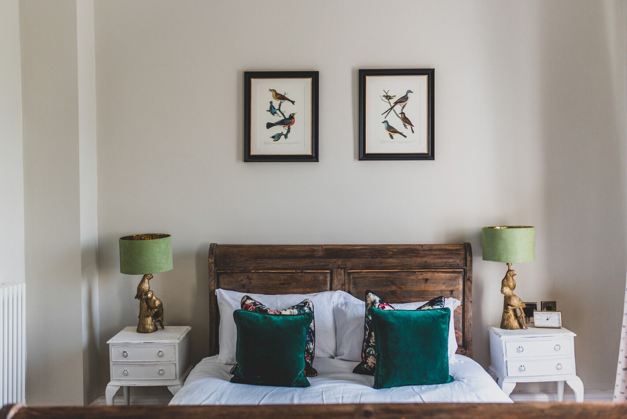 Une chambre à coucher avec une tête de lit en bois, deux illustrations d’oiseaux encadrées au mur, un lit avec des coussins verts et deux tables de chevet avec des lampes.