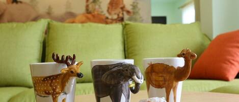 Three mugs with animal designs on a table in a room with green cushions and a red pillow.