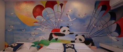 Habitación de hotel con dos camas, cada una con un almohadón que presenta un personaje de dibujos animados. La habitación tiene un mural colorido en la pared que representa a un panda y un globo aerostático.