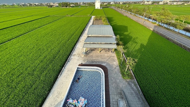 A large, rectangular swimming pool with a tiled deck area, surrounded by lush green fields and a road with a guardrail. - Wunuan ZOO Family Homestay (Jiaoxi)