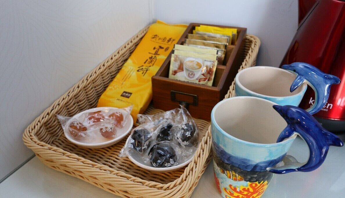 A wicker tray with packaged snacks, two ceramic mugs, and a red kettle.