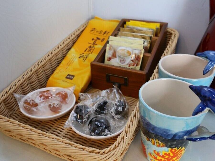 A wicker tray with packaged snacks, two ceramic mugs, and a red kettle.