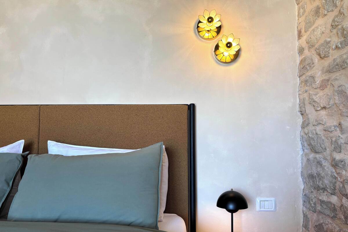 Ein Schlafzimmer mit braunem Kopfteil, einem Bett mit grün-weißer Bettwäsche, einem Nachttisch mit schwarzer Lampe und zwei Wandleuchten.