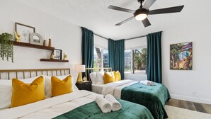 Habitación de hotel con dos camas, ventilador de techo, ventana con cortinas y repisa de pared con adornos.