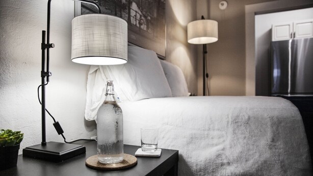Un lit bien fait, recouvert d’une couette blanche, une table de chevet avec une lampe, un verre d’eau et une bouteille d’eau.