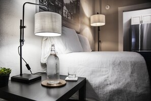 Una cama bien tendida con edredón blanco, una mesita de noche con lámpara, un vaso de agua y una botella de agua.