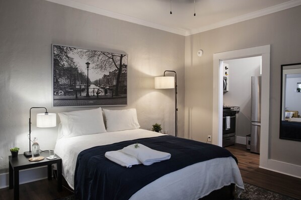 Ein modernes Hotelzimmer mit einem ordentlich bezogenen Bett, einem Nachttisch mit Lampe, einem Bild an der Wand und Blick in den Küchenbereich.
