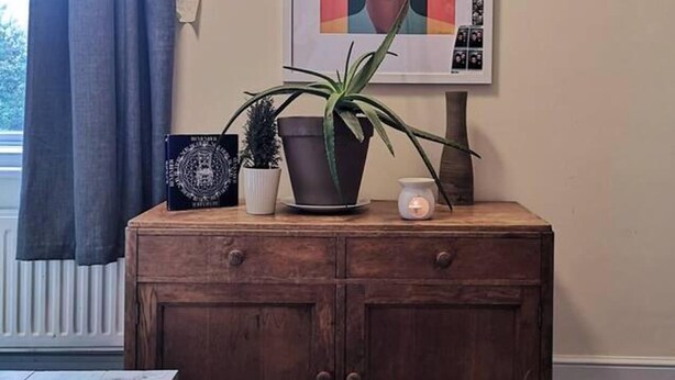 A wooden cabinet with a framed artwork, a potted plant, and a candle on top. - Cosy 1 Bed Flat Hideout in North London (London)