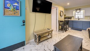 Ein Wohnzimmer mit Fernseher, einem Holztisch, einer blauen Tür und einer Küche im Hintergrund.