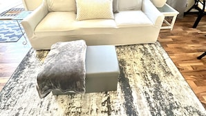 Un salon avec un canapé beige, un plaid gris et un tapis à motifs.