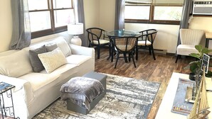 Un salon comprenant un canapé, un pouf, une table basse et une table à manger avec des chaises. De grandes fenêtres sont présentes, ornées de rideaux, ainsi qu’un ventilateur de plafond et une plante en pot sur la table basse.
