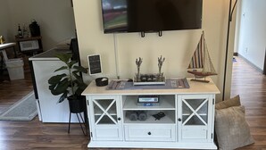 Un salon avec un téléviseur à écran plat fixé au mur, un meuble de rangement blanc sur lequel est posé un jeu d’échecs, une petite décoration représentant un voilier et une grande plante.