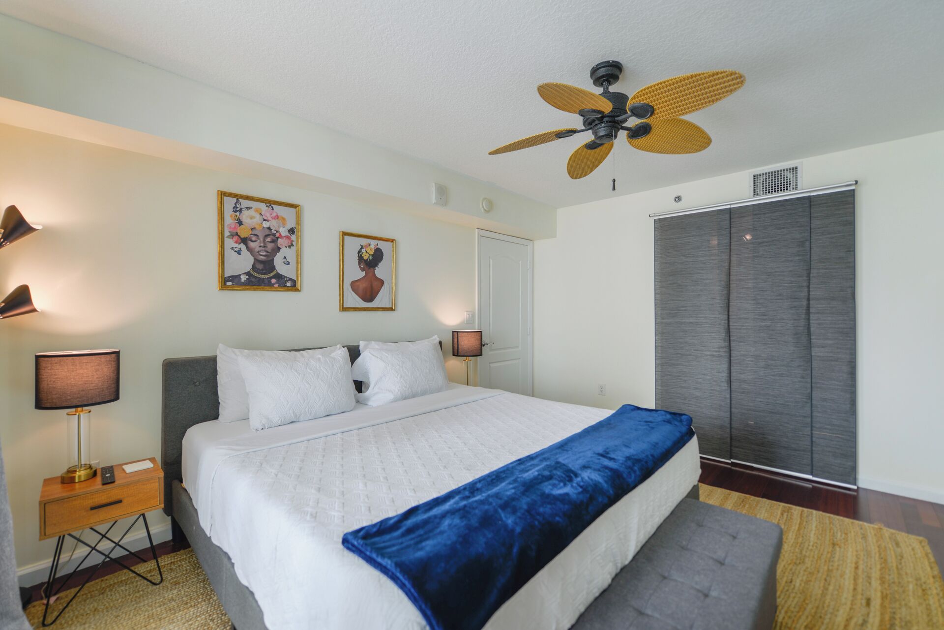 A bedroom with a bed, bedside lamps, a ceiling fan, a wardrobe, and two framed artworks on the wall.