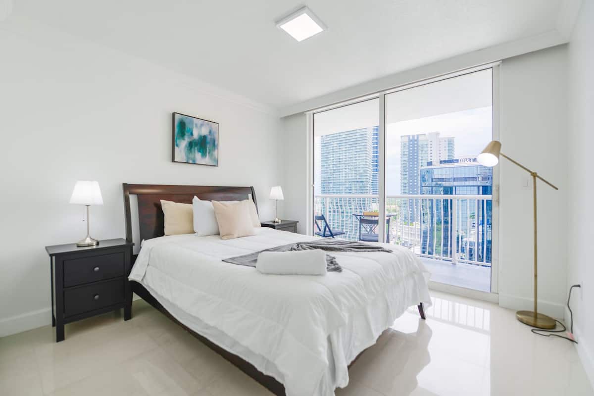 A modern bedroom with a large bed, two bedside tables, a nightstand, a floor lamp, and a city view through a large window.