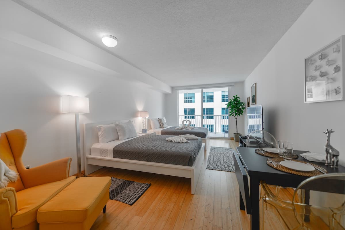 A modern hotel room with a bed, a desk, a chair, and a balcony with a view of buildings.