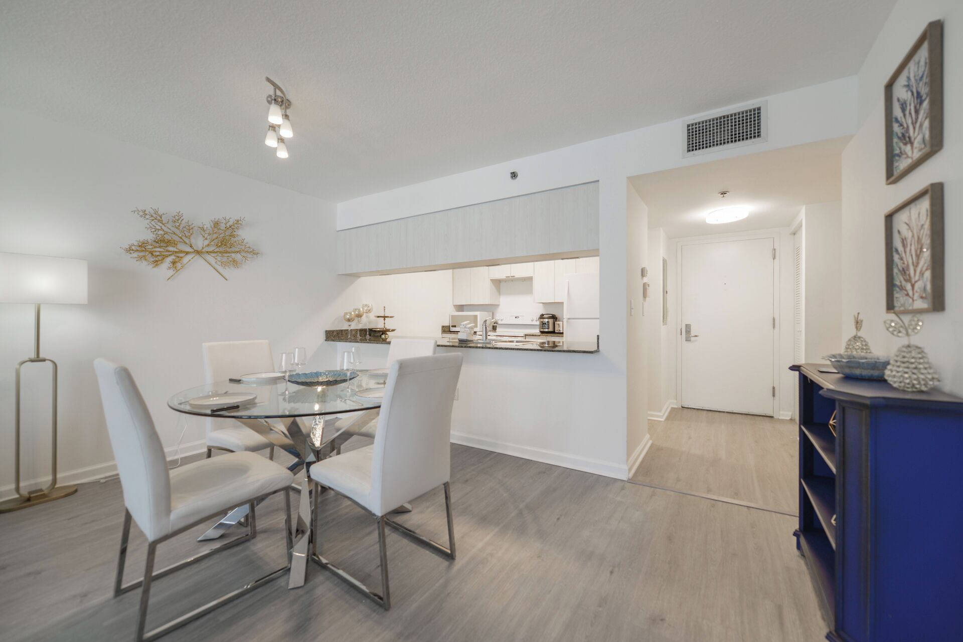 Una cocina moderna con armarios blancos, una zona de comedor con mesa de vidrio y sillas blancas, y un mueble azul con objetos decorativos.