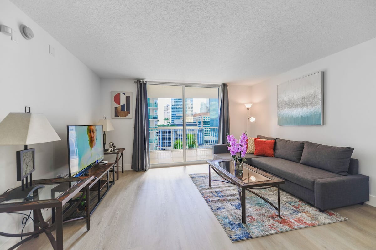 A modern living room with a grey sofa, a coffee table, a TV, and a large window with a city view.