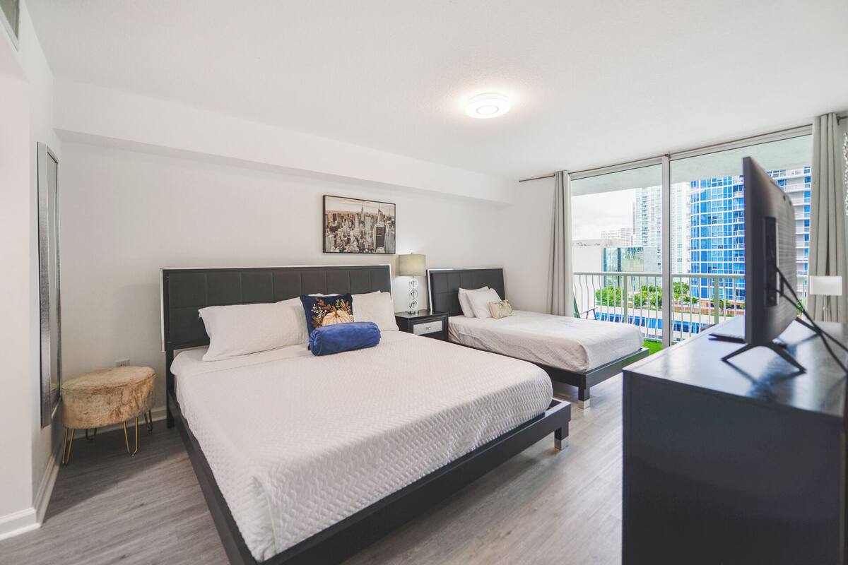A hotel room with two beds, a desk, a chair, a small stool, a nightstand, a lamp, a picture on the wall, and a city view through large windows.