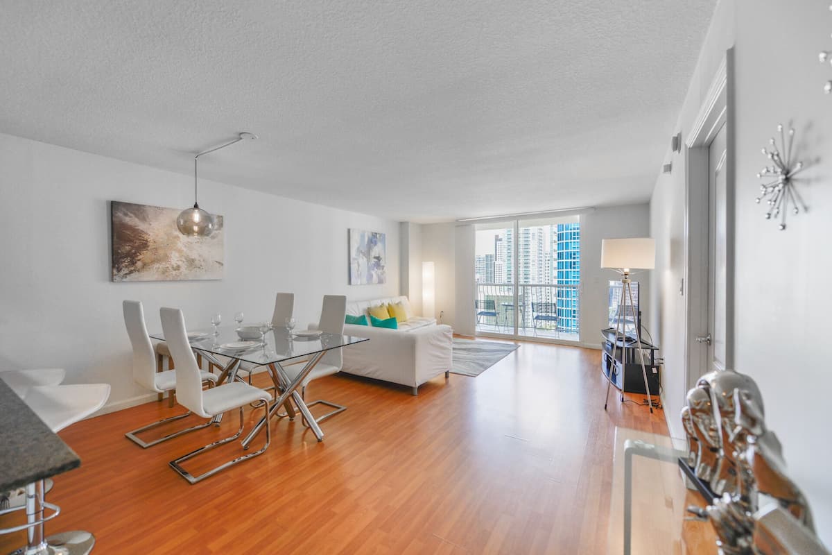 A spacious living area with a dining table, chairs, a sofa, and a large window offering a city view.