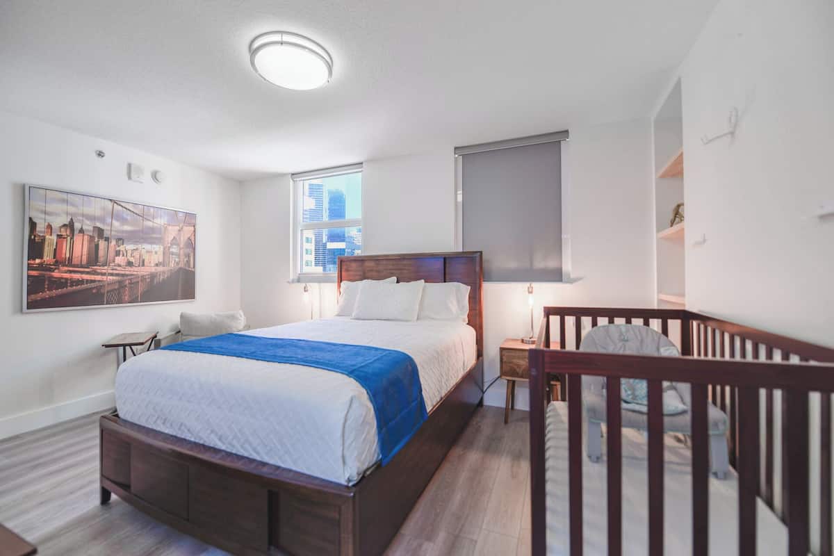 A bedroom with a bed, a crib, a cityscape painting, a window with a view, and a ceiling light.