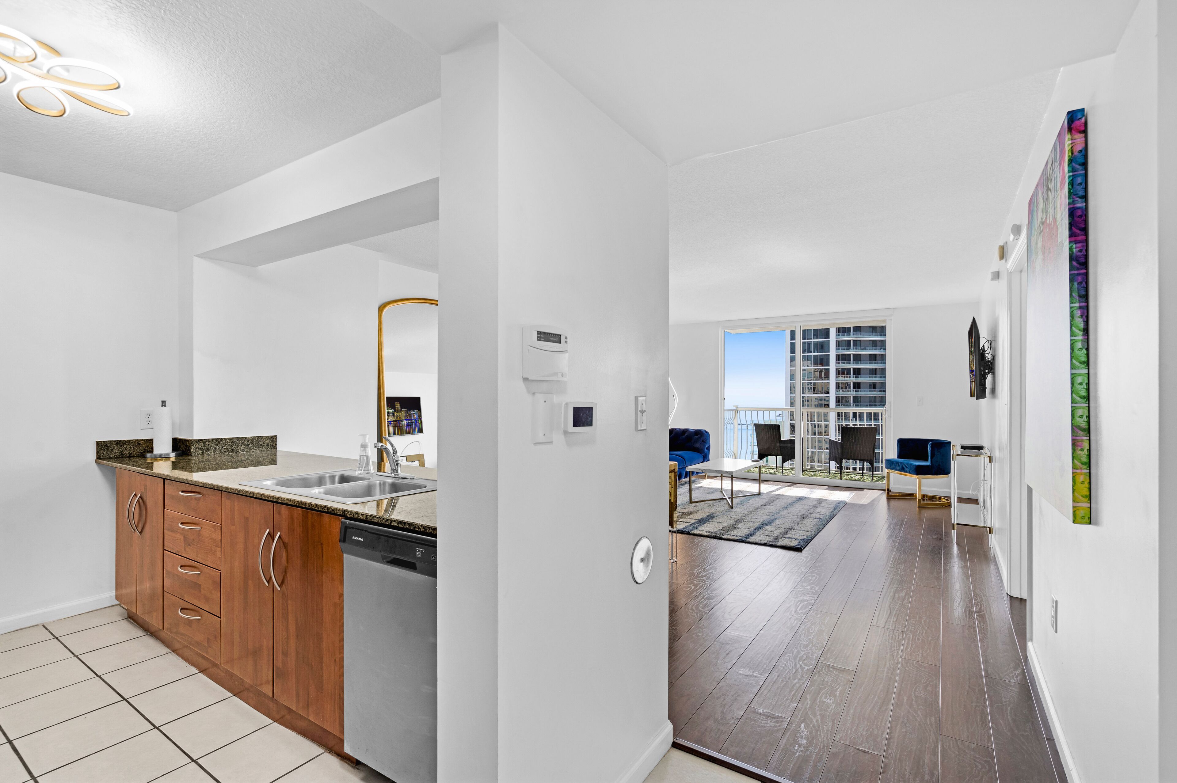 A modern kitchen with wooden cabinets and a dishwasher, a dining area with blue chairs, and a large window offering a view of the city.