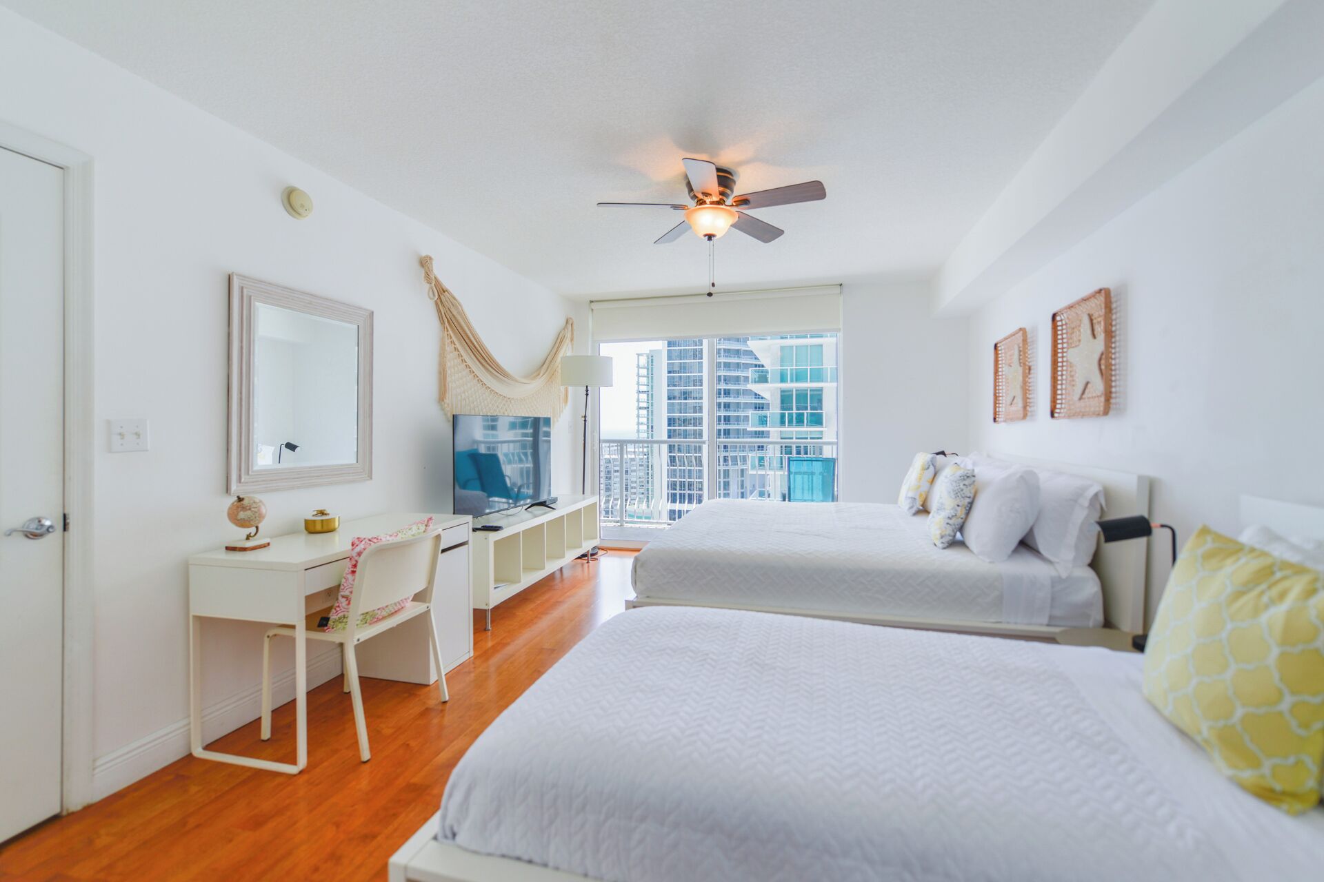 A modern bedroom with two beds, a desk, a chair, a ceiling fan, and a large window with a city view.