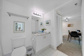 A modern bathroom with a toilet, vanity, and a mirror with lights. There are framed pictures on the wall and a small table with chairs in the adjacent room. - The Club at Brickell Bay (Miami)
