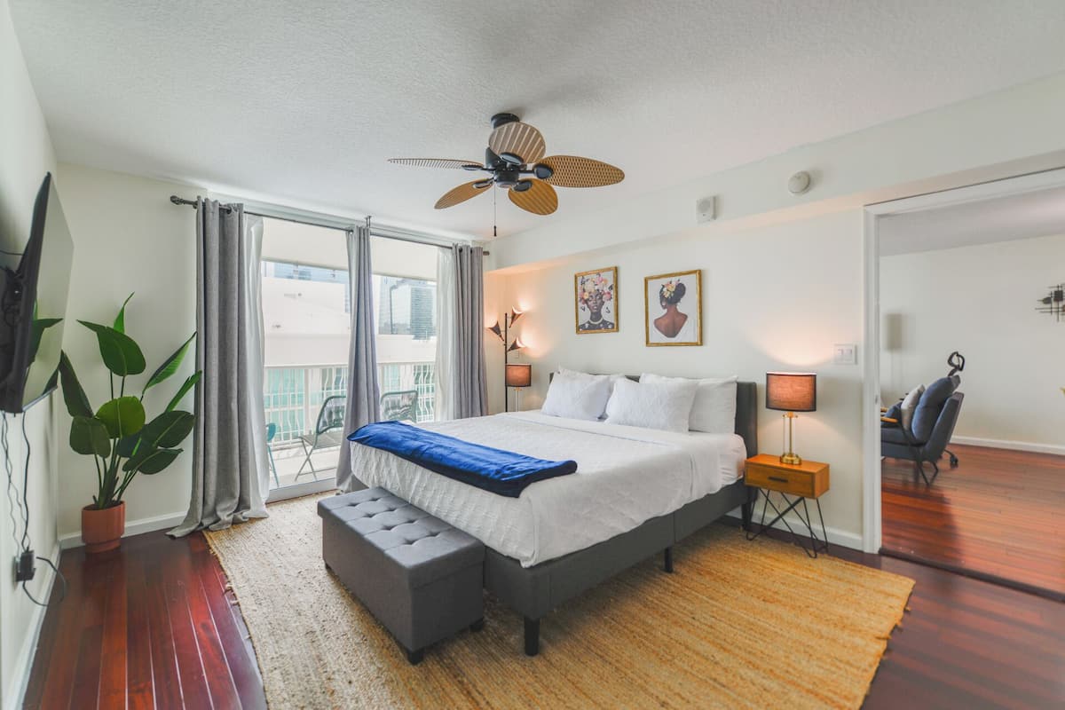 A bedroom with a bed, a bench, a ceiling fan, a nightstand, a lamp, a chair, and a balcony with a view.