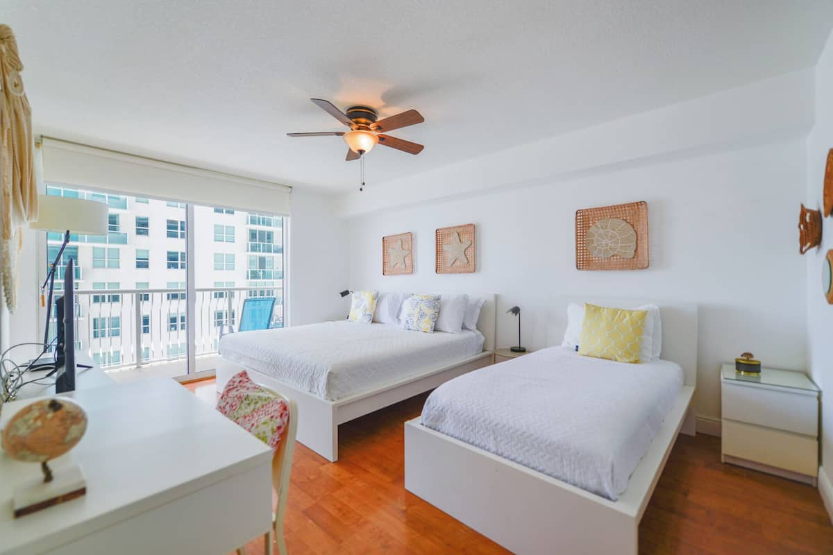 A bedroom with two beds, a ceiling fan, and a view of a building through large windows.