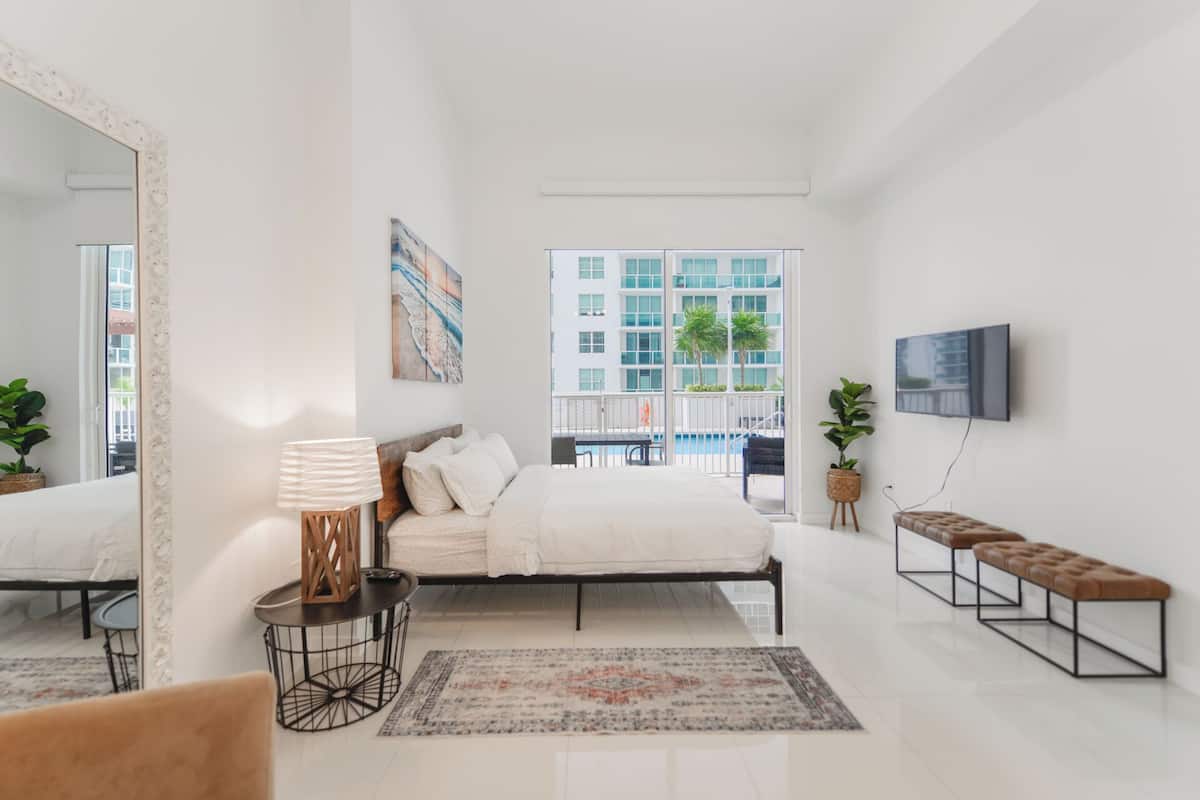 A modern bedroom with a large bed, a bedside table, a TV, a bench, and a view of a building through a window.